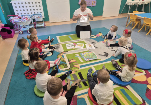 dzieci siedzą na dywanie, opiekunka pokazuje ilustracje z symbolami Polski