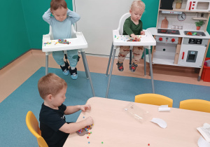 dzieci siedzą przy stoliku i w krzesełkach, wyklejają jeża kolorowymi kulkami, które są rozsypane na stole