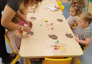 dzieci siedzą przy stoliku, a opiekunki pomagają im wykonać zadanie