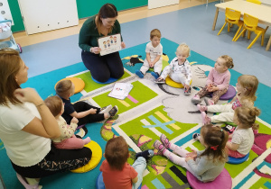 dzieci i opiekunki siedzą na dywanie, opiekunka pokazuje ilustracje