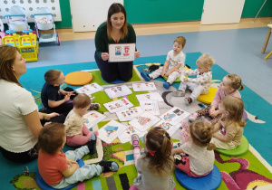 dzieci wraz z opiekunkami siedzą na dywanie, sluchają opiekunki i oglądają ilustracje