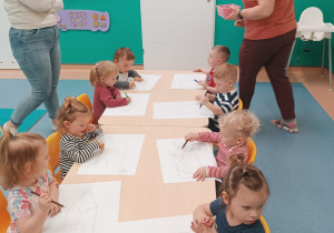 dzieci siedzą przy stoliku, opiekunka rozdaje dzieciom kredki