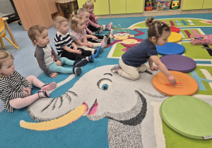 dzieci siedzą na dywanie, na dywanie leżą kolorowe kółka dziewczynka kładzie kredkę na kółku w takim samym kolorze