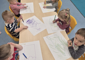 dzieci siedzą przy stoliku i kolorują narysowane na kartonie kredki