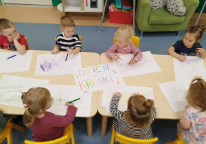 dzieci przy stoliku , na kartonach kolorują kredkami narysowaną dużą kredkę