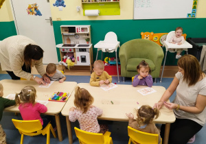 dzieci siedzą przy stoliku i wykonują zadanie , opiekunki pomagają