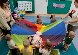 dzieci i opiekunki trzymają kolorową chustę na której leżą pluszowe misie