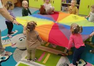 dzieci trzymają kolorową chustę i podrzucają leżące na niej misie