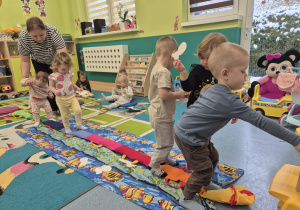 dzieci zbierają rybki z podłogi, wkładają je do pyszczka misia, opiekunka podtrzymuje małe dziecko