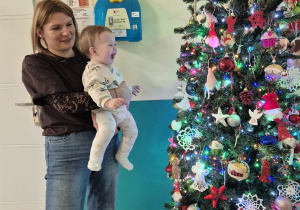opiekunka trzyma na rękach dziecko, dziecko uśmiecha się na widok choinki