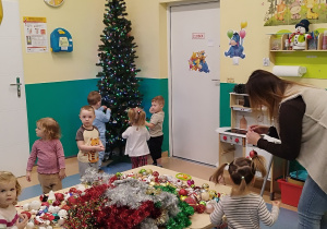 opiekunka pomaga dziewczynce z bombką, inne dzieci wieszają je na choince