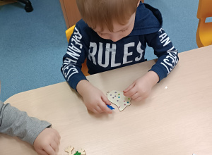 dzieci siedzą przy stoliku i przyklejają dekoracyjne diamenciki do drewnianej bombki