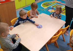 dzieci siedzą przy stoliku wyklejają diamencikami swoje kształty, chłopiec sięga talerzyk z ozdobami