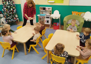 dzieci siedzą przy stolikach, opiekunka rozdaje im drewniane kształty