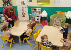 dzieci siedzą przy stolikach, opiekunka rozdaje im drewniane kształty