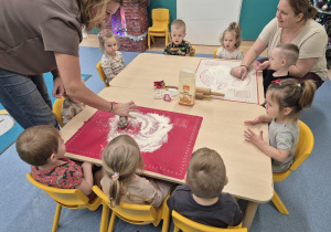 dzieci siedzą i patrzą, a opiekunki pokazują jak ugniatać ciasto