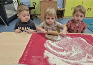 dziewczynka siedzi między dwoma chłopcami i wałkuje ciasto