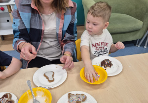 chłopiec bierze posypkę i patrzy jak obok opiekunka dekoruje pierniczka