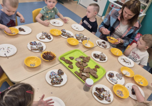 dzieci przy stoliku dekorują pierniczki, opiekunka pomaga