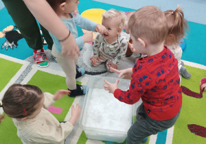 dzieci dotykają śniegu w pojemniku, opiekunka pomaga chłopcu