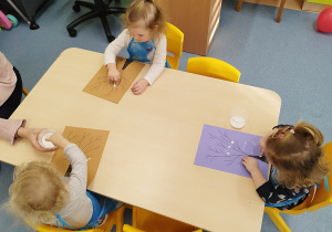 dzieci siedzą przy stoliku, przed nimi leżą kartki, na których malują, opiekunka pomaga