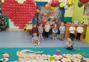 dzieci stoją i siedzą w tle sylwetki babci i dziadka,chłopiec siedzi na dywanie bawi się balonem