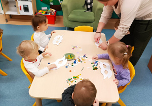 opiekunka smaruje maskę klejem, dzieci siedzą przy stoliku