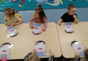 dzieci siedzą przy stoliku trzymając szczoteczki, przed nimi leżą buźki do wyczyszczenia
