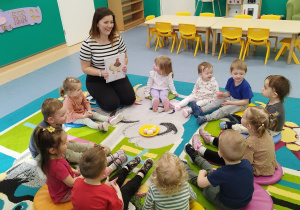 Dzieci siedzą z opiekunką na dywanie, opiekunka pokazuje im obrazek z kurką