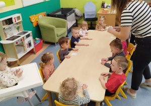 dzieci siedzą przy stoliku, opiekunka rozdaje im elementy do pracy plastycznej