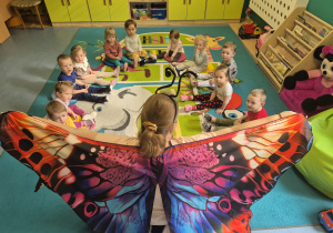 dzieci siedzą na dywanie, opiekunka pokazuje kolorowe skrzydła, które założyła