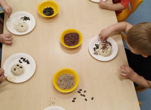 dzieci siedzą przy stoliku , na talerzykach mają ciasto, w które wkładają pestki dyni, słonecznika