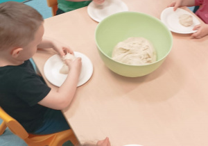 dzieci siedzą przy stoliku, przed nimi leżą talerzyki z kulką ciasta