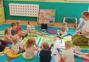 opiekunki wraz z dziećmi siedzą na dywanie , opiekunka pokazuje ilustracje