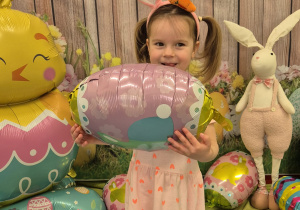 dziewczynka trzyma pisankę-balona, na głowie ma opaskę uszy królika, uśmiecha się
