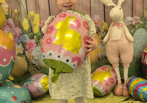 dziewczynka uśmiecha się, trzyma balona-pisankę, na głowie ma opaskę uszy królika