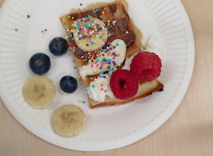 na talerzyku leży gofr z bitą śmietaną, z kremem czekoladowym, bananami, borówkami i maliną, posypany kolorową posypką