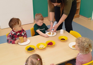 dzieci siedzą przy stoliku, opiekunka wyciska śmietanę na gofra