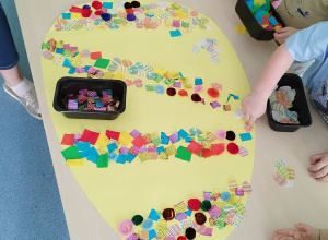 dzieci stoją wokół stolika i przyklejają ozdoby na papierowe jajko