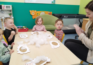 dzieci siedzą przy stoliku i wyklejają baranki razem z opiekunką
