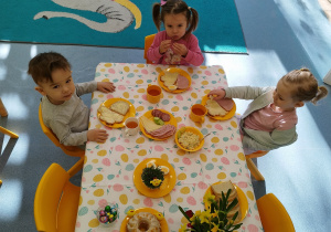 dzieci siedzą przy stole , chłopiec i dziewczynka patrzy w aparat, a druga dziewczynka sięga kanapkę z talerza