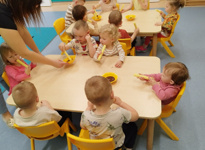 dzieci siedzą przy stoliku, trzymają chrupki, opiekunka podaje miseczkę z czekoladą