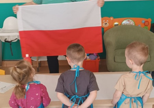 opiekunka pokazuje dzieciom plagę Polski, dzieci w fartuszkach stoją prze stoliku