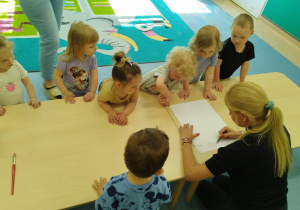 dzieci stoją wokół stolika, a pani pedagog rysuje na kartonie pień drzewa