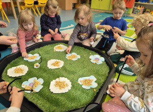 dzieci siedzą wokół pola , które jest łąką i nawlekają kółeczka na druciki