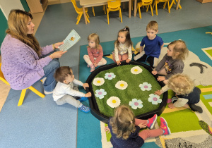 opiekunka czyta dzieciom, które siedzą wokół pojemnika z łąką