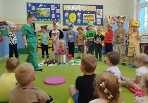 żlobkowe maluchy siedzą na dywanie, przed nimi stoją poprzebierane przedszkolaki i dziewczynka siedzi w wielkim garnku