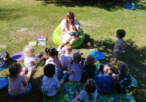 opiekunka siedzi na pufie i czyta książkę dzieciom siedzącym na przeciwko niej na kocu