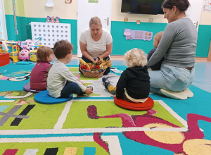 dzieci wraz z opiekunkami siedzą na dywanie, przed nimi stoi koszyk pełen warzyw i owoców, liści