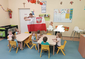 dzieci siedzą przy stoliku , opiekunka pokazuje im flagę Polski